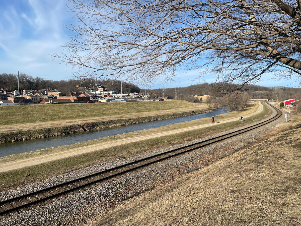 Rail lines and walking paths follow the Galena River beside the town’s historic center. Scenic landscape in Grant Park, Galena, Illinois showing a curving railway beside a narrow river, two cyclists riding along a dirt trail, and the town’s historic buildings in the background on a clear early‑spring day.