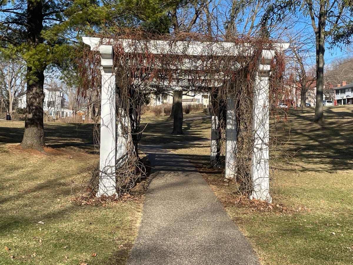 A white pergola overlooks the river valley from Grant Park above downtown. White pergola with dormant vines over a paved walkway in Grant Park, Galena, Illinois, surrounded by leafless winter trees and nearby homes.