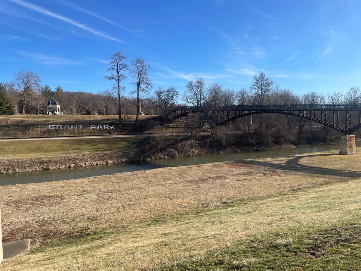 From Grant Park, the Galena River winds past the base of the historic downtown district. View of the arched metal bridge crossing the Galena River in Grant Park, Galena, Illinois, with “Grant Park” spelled out on the hillside, leafless trees, and a small green-roof gazebo under a clear blue sky.