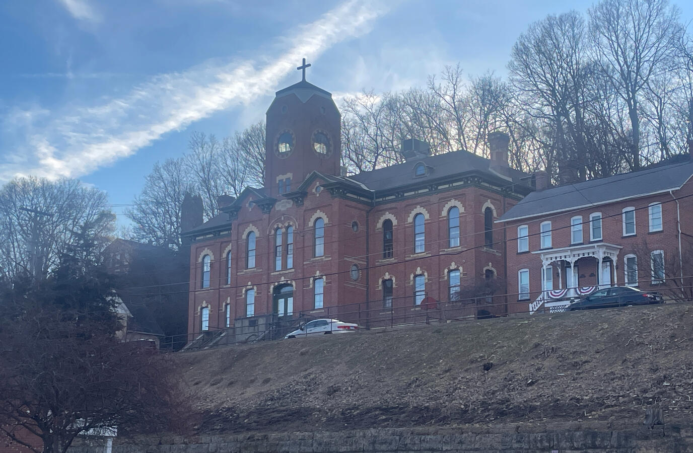 Brick commercial blocks rise along the hillside streets surrounding downtown. Historic 19th‑century brick building with a central tower and arched windows on a hilltop in Galena, Illinois, photographed in winter with leafless trees, parked cars, and a nearby brick home under a partly cloudy sky.